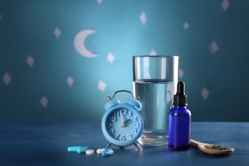 Deprived of Sleep, Many Turn to Melatonin Despite Risks Alarm clock and different remedies for insomnia treatment near glass of water on table against blue wall decorated with stars and crescent